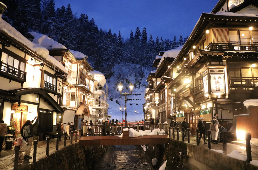 A picturesque winter scene in Ginzan Onsen town. (hot springs) Rows of beautifully lit wooden buildings line the snow-covered streets, with a small stream flowing through the center.