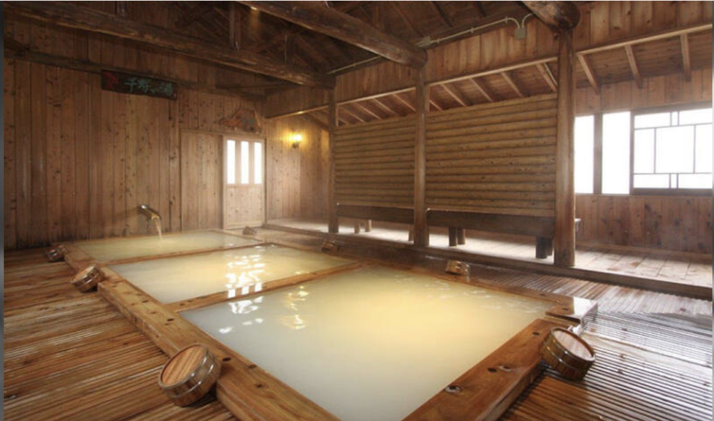 A traditional Japanese wooden bathhouse with two large hot spring baths. The baths are filled with steaming water and surrounded by wooden walls and benches.